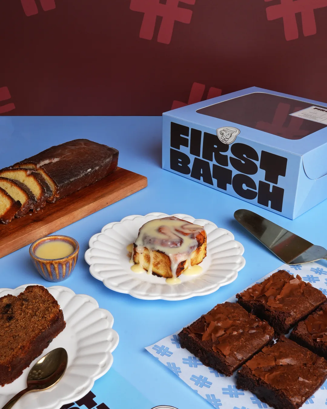 A First Batch spread on a # wall: banana bread, a frosted cinnamon roll, brownies and a First Batch box